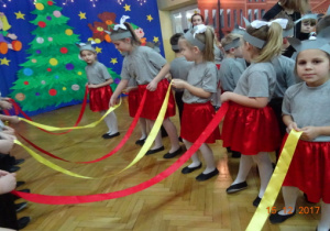 Dziewczynki z szarymi opaskami na głowach stoją w rzędzie naprzeciweko chłopców i trzymają żółte i czerwone wstążki.